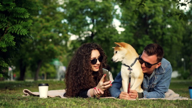 Junge-Frau-zeigt-lustige-Bilder-auf-Smartphone-zu-ihrem-Freund-und-entspannen-Sie-im-Park-mit-Hund,-Menschen-liegen-auf-Plaid-Gras-und-beobachten-Bildschirm.