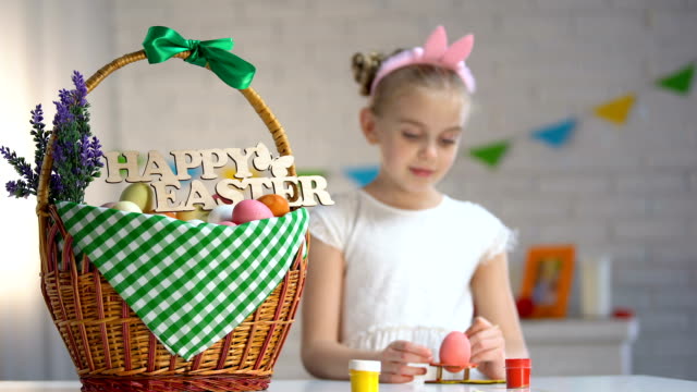 Happy-Easter-sign-on-basket,-cute-girl-painting-eggs-sitting-at-table,-holiday