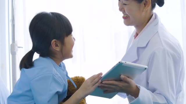 Female-doctor-showing-digital-tablet-computer-and-talking-with-young-patient-girl-with-happy-emotion.