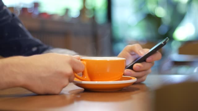 Nahaufnahme-der-Hand-eines-Mannes-halten-und-mittels-Smartphone-beim-Trinken-aus-einer-orangefarbenen-Tasse-dampfenden-Kaffee-in-ein-modernes-Café-in-Medellin,-Kolumbien,-Lateinamerika.