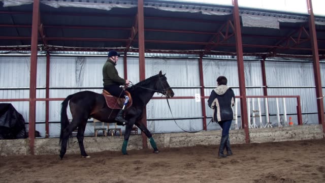 in-special-hangar,-a-young-disabled-man-learns-to-ride-a-horse-with-close-supervision-teacher,-hippotherapy.-man-has-artificial-limb-instead-of-his-right-leg.-rehabilitation-of-disabled-with-animals