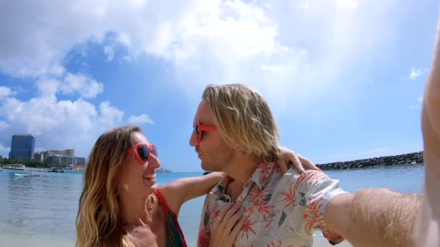 Couple-taking-selfies-on-Waikiki-Beach-in-Honolulu,-Hawaii.-Selfie-point-of-view-wide-angle-of-Waikiki-Beach.-Young-couple-taking-selfies-with-heart-shaped-sunglasses