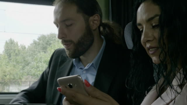Closeup-of-young-business-partners-couple-sitting-in-a-moving-train-talking-and-analyzing-business-information-on-smartphone