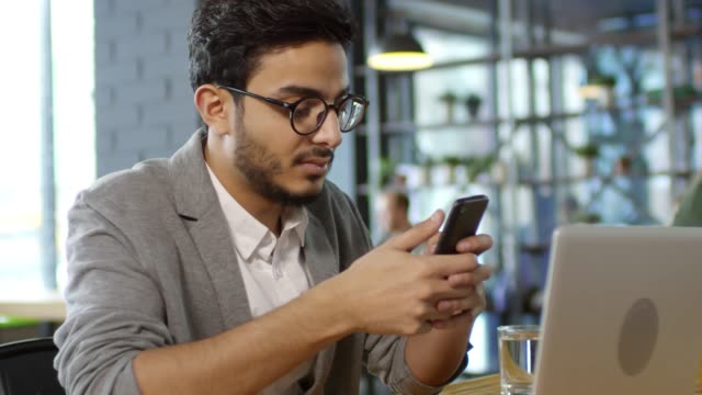 Hübsche-arabische-Mann-mit-Smartphone-und-Lächeln-bei-Kamera-im-Cafe