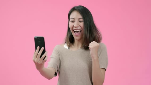 Junge-asiatische-Frau,-die-Smartphone-überprüft-Social-Media-Gefühl-glücklich-lächelnd-in-lässiger-Kleidung-über-rosa-Hintergrund-Studioaufnahme-erschossen.-Glücklich-lächelnde-entzückende-Frau-freut-sich-über-Erfolg.