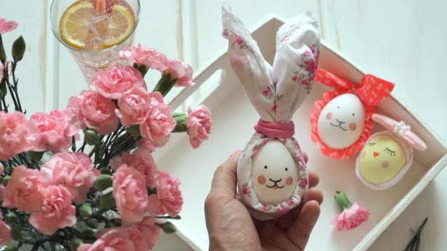Hand-holds-Easter-egg-decorated-for-Easter-bunny-with-ears-and-painted-muzzle,-against-background-of-decorated-eggs,-pink-carnations-and-lemonade