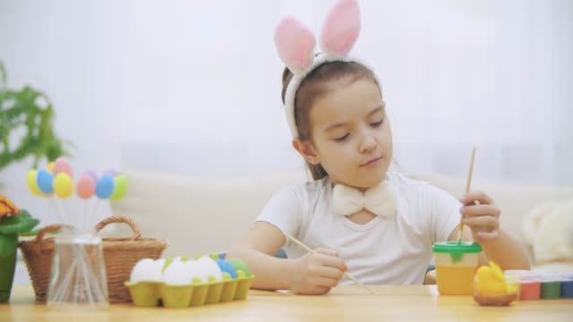 Little-cute-girl-is-having-fun-holding-a-paint-brush-in-her-left-hand.-Girl-with-a-beauty-spots-at-her-face-watching-at-nude-paint-brushes,-sitting-at-the-wooden-table-with-Easter-decorations.-Girl-is-amused-and-gives-up-holding-paint-brushes-and-hiding-h