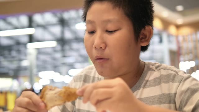 Happy-Asian-preteen-boy-eating-fried-chicken-leg-in-fast-food-restaurant.