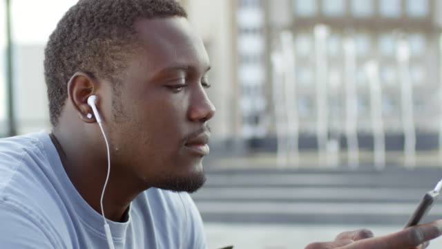 Hombre-africano-feliz-escuchando-música-en-el-teléfono-inteligente-y-sonriendo-a-la-cámara
