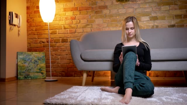 Closeup-portrait-of-young-pretty-blonde-haired-female-using-the-tablet-sitting-on-the-floor-carpet-indoors-in-a-cozy-apartment