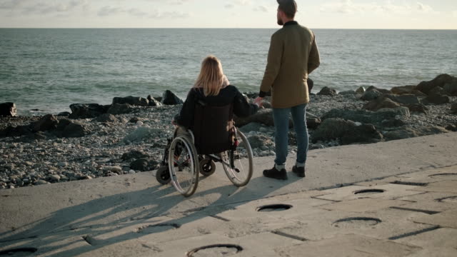 Woman-is-sitting-in-wheelchair-on-sea-coast-and-holding-hand-of-her-husband