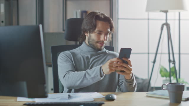 Handsome-Long-Haired-Entrepreneur-Sitting-at-His-Desk-in-the-Office-Works-on-Desktop-Computer,-Working-with-Documents,-Graphs.-Uses-Smartphone,-Social-Media-App,-Writing-Emails,-Messaging