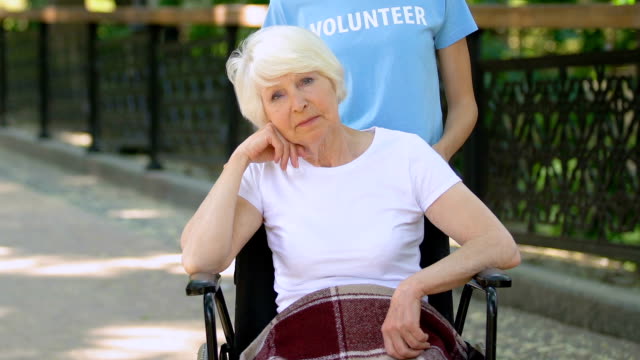 Volunteer-with-upset-senior-woman-in-wheelchair-looking-at-camera,-hospital-park