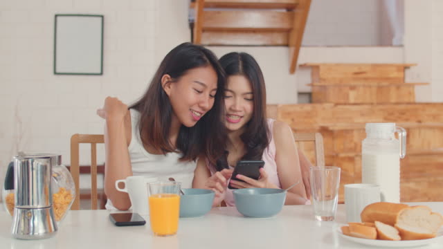 Asian-Lesbian-couple-using-mobile-phone-check-news-while-has-breakfast-in-kitchen-at-home.