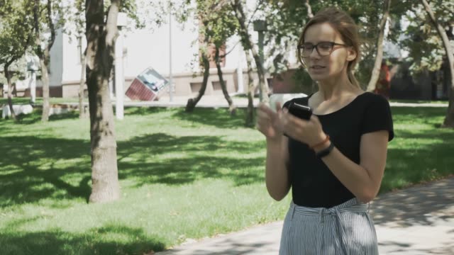 Hübsche-junge-kaukasische-Frau-in-Brille-zu-Fuß-in-Richtung-Kamera,-Überprüfen-DesTelefon-und-feiern-gute-Nachrichten-in-einem-Park