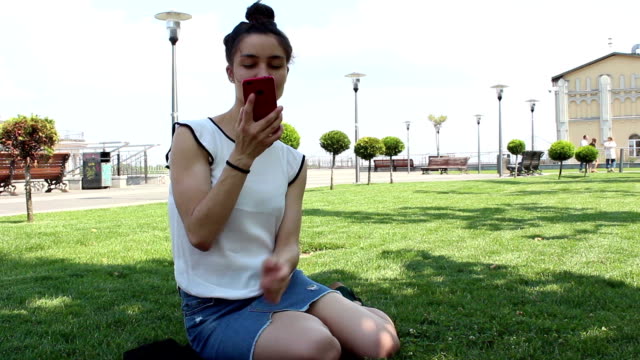A-young-attractive-girl-in-a-white-T-shirt-is-smiling-and-looking-through-the-tape-of-social-networks-in-her-smartphone-while-sitting-on-the-grass-in-the-park.