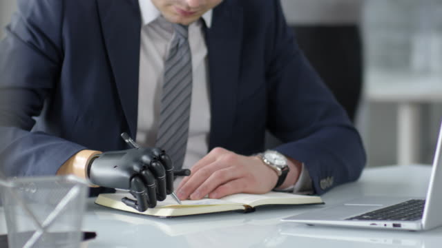 Businessman-with-Bionic-Hand-Writing-in-Notepad-at-Desk