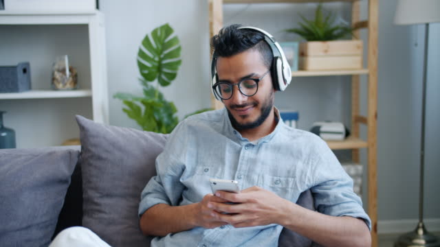 Guapo-árabe-chico-usando-teléfono-inteligente-escuchando-música-en-auriculares-en-casa