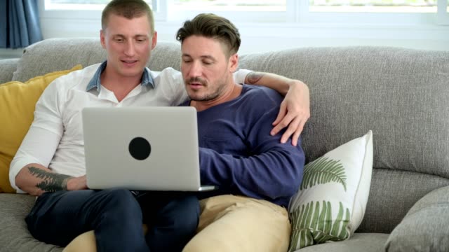 Pareja-gay-relajándose-en-el-sofá-usando-la-computadora-portátil.-Completar-la-transacción.