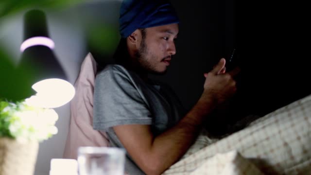 Attractive-Asian-man-using-a-mobile-phone-while-Lying-on-Bed-at-home.-Browsing-social-media-and-watching-a-video.