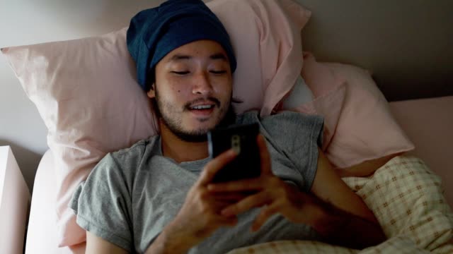 Schließen-Sie-hübsche-asiatische-Mann-mit-einem-Mobiltelefon,-während-liegen-auf-dem-Bett-zu-Hause-Late-at-Night.-Durchsuchen-von-sozialen-Medien-und-Ansehen-eines-Videos.