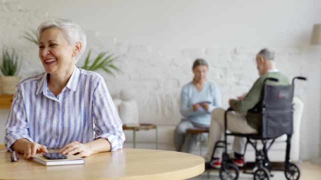 Tracking-Aufnahme-von-älteren-Paar-am-Tisch-sitzen-und-Spaß-im-Gespräch-freudig,-Seniorin-und-behinderte-Senior-In-Rollstuhl-Spielkarten-im-Hintergrund-in-betreutem-Wohnen-zu-Hause