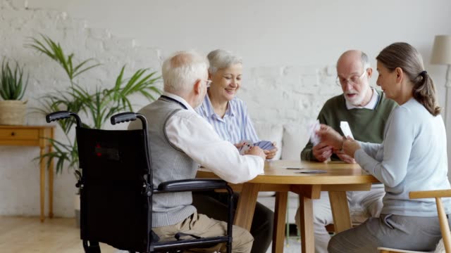 Tracking-shot-of-group-of-four-retired-elderly-people,-two-men-and-two-women,-sitting-at-table-and-playing-cards-together-in-common-room-of-assisted-living-home