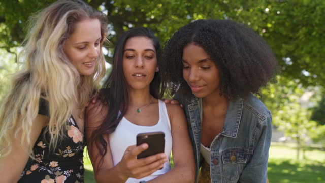Glückliche-vielfältige-junge-Freundinnen-beobachten-Inhalte-auf-dem-Smartphone-Bildschirm-im-Park---Freunde-im-Park-mit-einer-mobilen-App