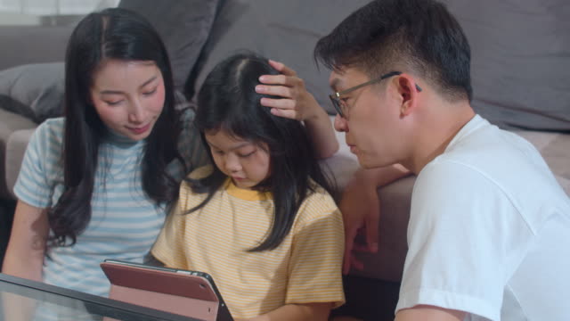 Young-Asian-family-and-daughter-happy-using-tablet-at-home.-Japanese-mother,-father-relax-with-little-girl-watching-movie-lying-on-sofa-in-living-room.-Funny-parent-and-lovely-child-are-having-fun.