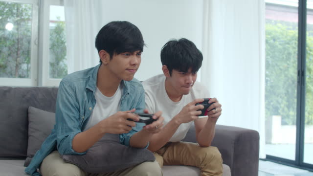 Joven-pareja-gay-asiática-jugar-juegos-en-casa,-adolescentes-coreanos-HOMBREs-LGBTQ-usando-joystick-que-tienen-divertido-momento-feliz-juntos-en-el-sofá-en-la-sala-de-estar-en-casa.