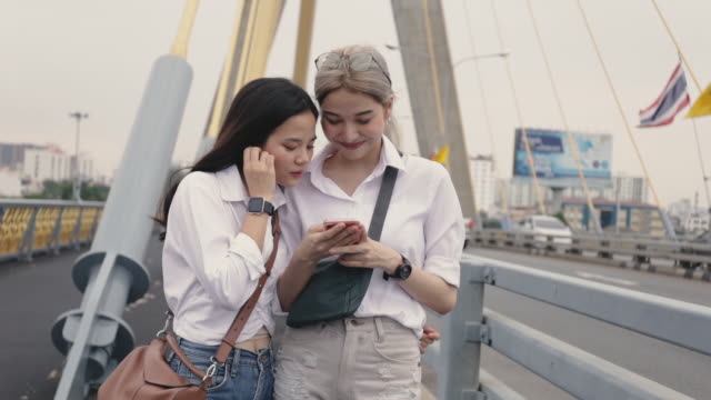 Asiatische-lesbische-Paare,-die-Smartphones-nutzen,-um-auf-der-Website-zu-surfen,-während-sie-auf-der-Brücke-stehen.-Low-Winkel-Schuss.