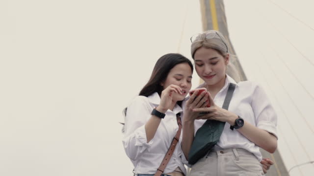 Asiatische-lesbische-Paare,-die-Smartphones-nutzen,-um-auf-der-Website-zu-surfen,-während-sie-auf-der-Brücke-stehen.-Low-Winkel-Schuss.