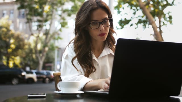Female-in-glasses,-white-shirt.-She-sitting-at-table-with-laptop-and-smartphone-in-a-roadside-cafe,-drinking-coffee-and-working-online.-Close-up