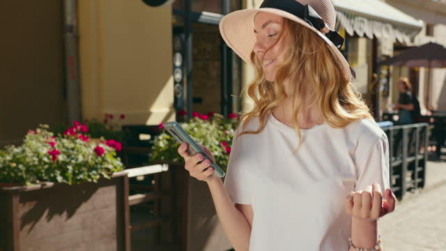 Ein-Mädchen-geht-durch-die-Stadt-und-sms-auf-ihrem-Smartphone.-Einkaufstaschen-hängen-an-ihrem-Arm.-Sie-trägt-einen-Hut-und-eine-Sonnenbrille.-4K