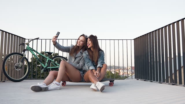 Ziemlich-jovial-sexy-Mädchen-sitzen-auf-dem-Skateboard-und-machen-Selfie-mit-lustigen-Gesichtern-auf-der-Dachterrasse