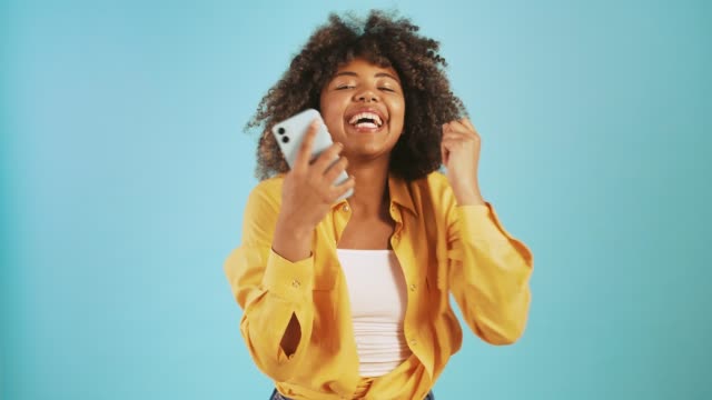Afro-american-lady-looking-at-smartphone,-saying-yes,-raising-fists-up-being-surprised-and-overjoyed,-smiling-posing-on-blue-background