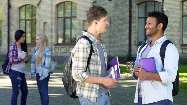 Two-smiling-male-students-talking-about-graduation-party-near-university
