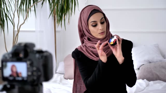 Young-beautiful-indian-girl-in-hijab-blogger-talking-on-camera,-smiling,-showing-a-new-purchase,-cosmetics,-aquagrum,-home-comfort-in-the-background.-60-fps