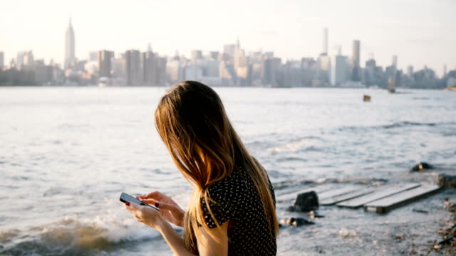 Nicht-erkennbare-Frau-mit-langen-Haaren-mit-Smartphone-app-am-Flussstrand-mit-New-York-City-Skyline-Blick-auf-Sonnenuntergang-4K