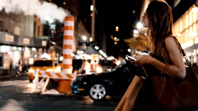 Joven-hermosa-mujer-cruzando-la-carretera-de-tráfico-en-el-centro-de-Nueva-York,-América-en-la-noche-y-el-uso-de-smartphone