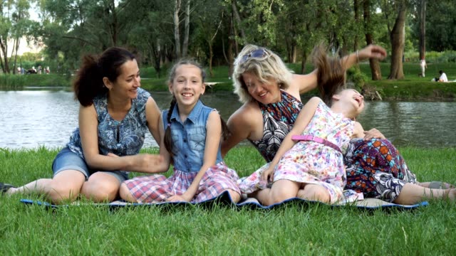 Homo-Familie:-zwei-Mütter-mit-zwei-Töchter-spielen,-lachen-und-Spaß-haben,-sitzen-im-Park-auf-dem-Rasen-bei-Sonnenuntergang-in-der-Nähe-des-Flusses.
