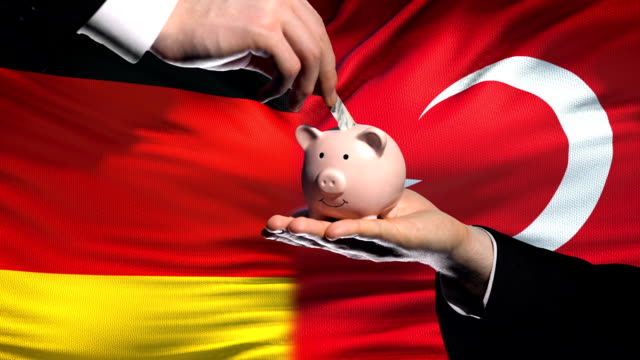 Germany-investment-in-Turkey,-hand-putting-money-in-piggybank-on-flag-background