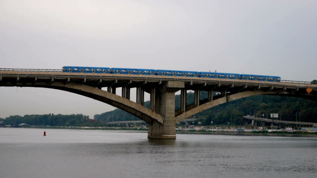Train-travels-over-the-bridge-against-the-sky-and-forest.-City-subway-left-outside.-Public-transport-on-the-move.-Cars-go-over-the-bridge-over-the-river.