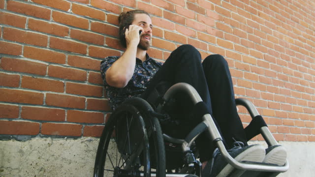 Joven-discapacitado-realizar-llamada-telefónica-en-silla-de-ruedas-inclinada-contra-la-pared-de-ladrillo