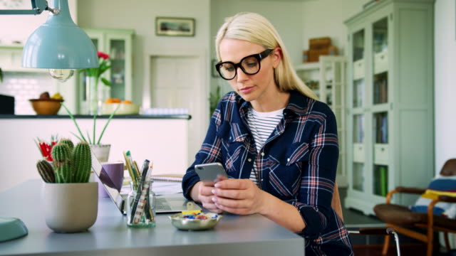 Attraktive-blonde-Geschäftsfrau-SMS-auf-Smartphone-im-Home-Office