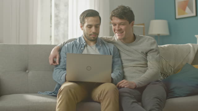 Entzückende-männliche-Gay-paar-verbringen-Zeit-zu-Hause.-Sie-sitzen-auf-einem-Sofa-und-das-Notebook-nutzen.-Stöbern-sie-Online.-Partners-Hand-ist-um-seine-Geliebte.-Zimmer-ist-Modern-eingerichtet.