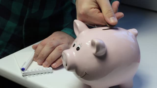On-the-table-there-is-a-piggy-bank-in-the-form-of-a-pink-pig.-A-man-throws-coins-at-her.-Makes-notes-in-a-notebook.