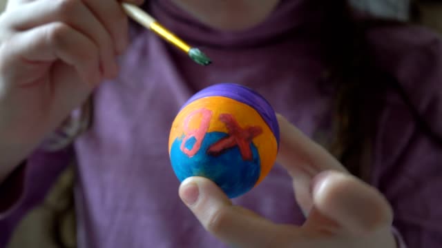 A-little-girl-holds-an-Easter-egg-in-her-hand-and-paints-on-it-a-brush-and-colored-paints,-sitting-at-home-at-the-table.-Close-up.-4K.