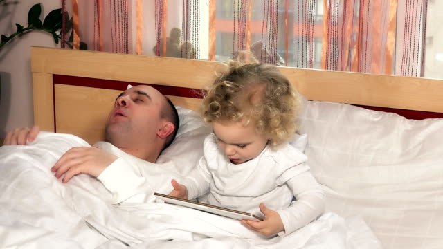 bald-dad-fall-asleep-while-daughter-girl-play-with-tablet-computer-sit-in-bed