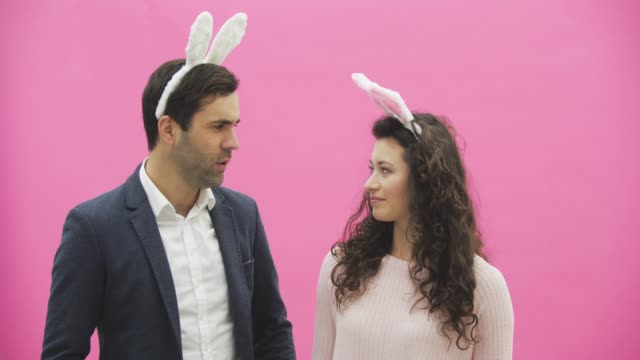 Beautiful-young-couple-standing-on-pink-background.-At-the-same-time,-looking-at-the-camera,-they-smile.-Together-with-pink-barking-ears-on-the-head.-Happy-family-is-preparing-for-Easter.